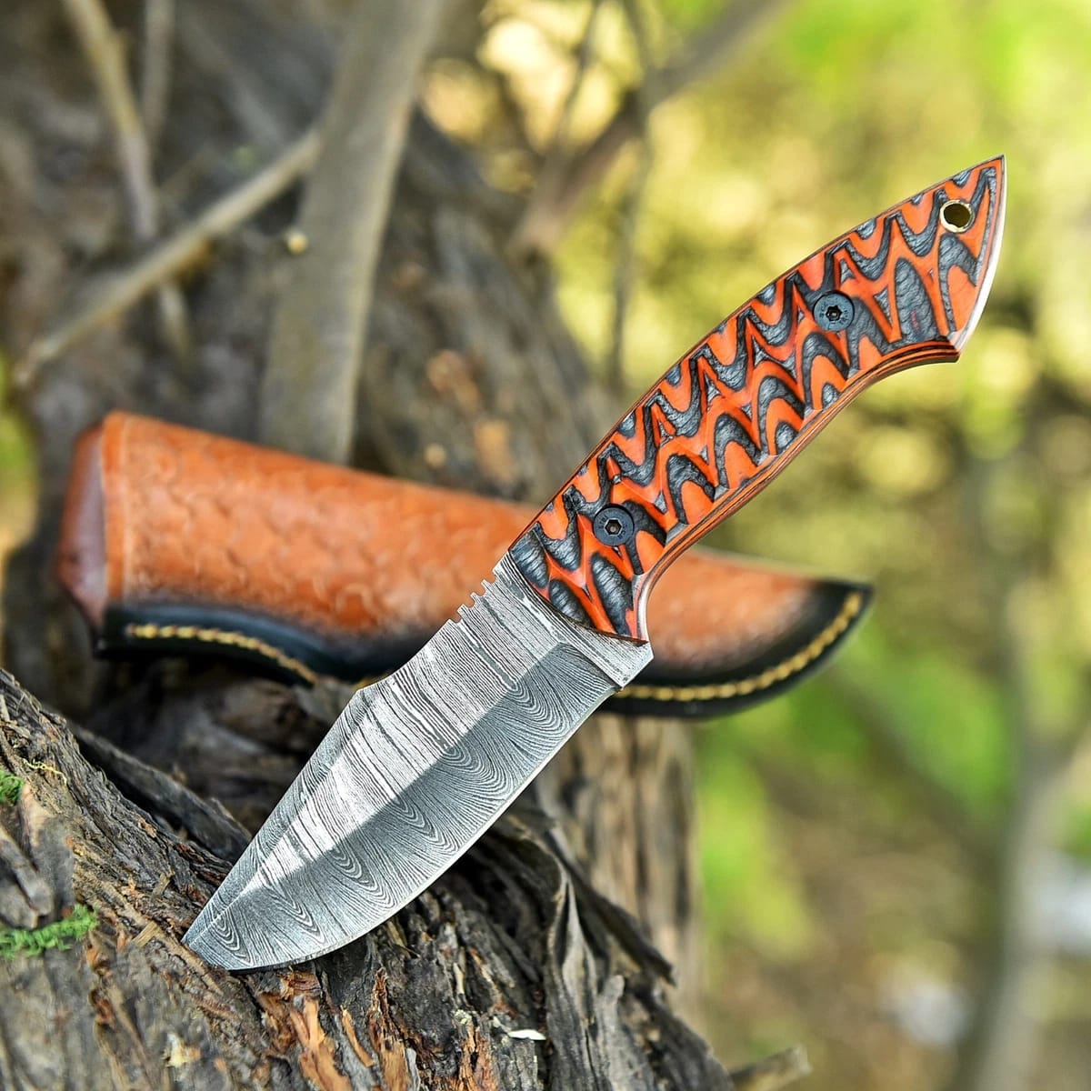 Hand-forged Damascus steel fixed blade knife with textured orange and black G10 handle resting on a tree branch next to a basketweave leather sheath