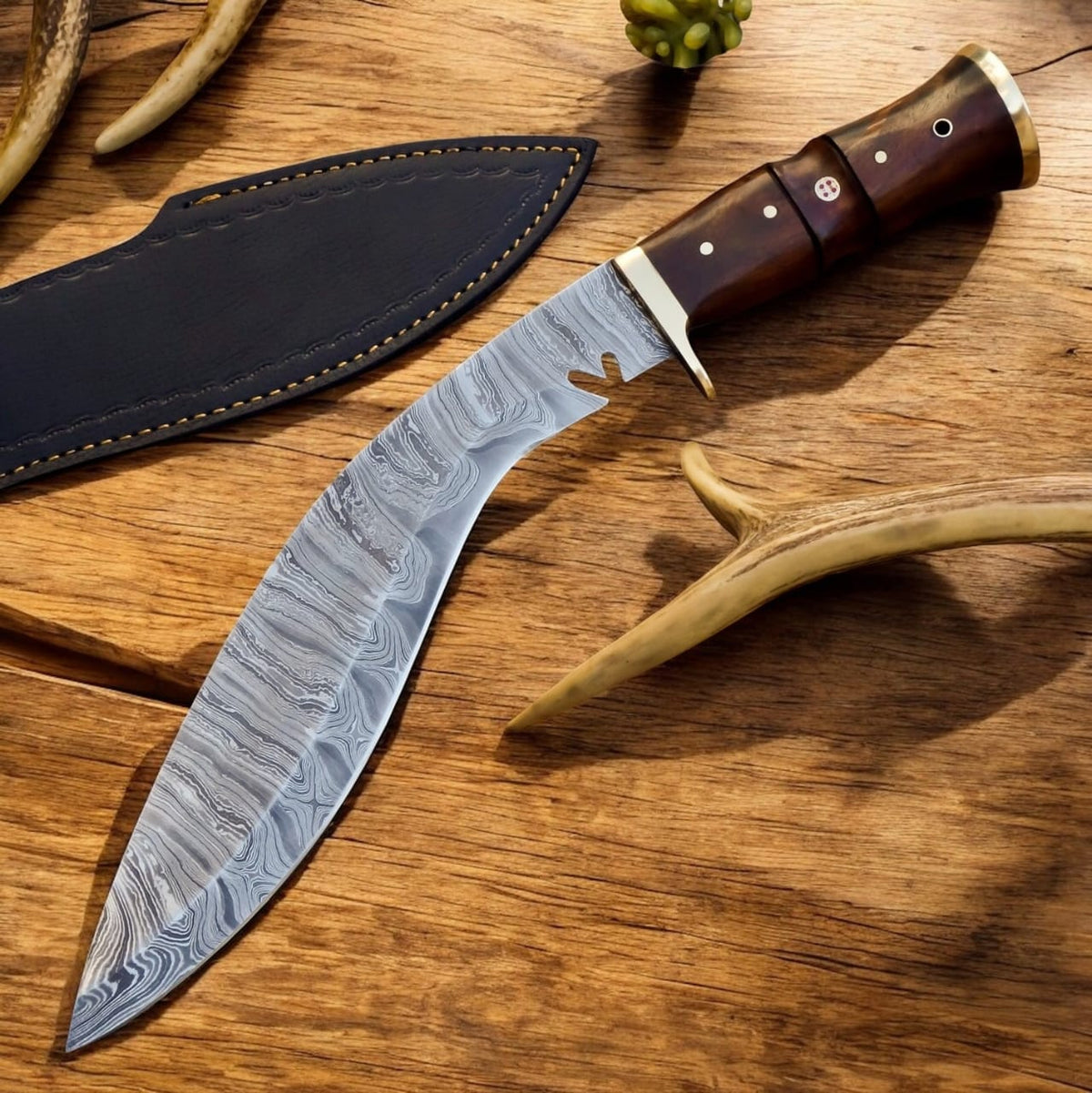 A curved Damascus steel knife with a patterned steel blade, dark brown wooden handle, and brass bolsters, displayed next to its black leather sheath on a rustic wooden surface with antlers.