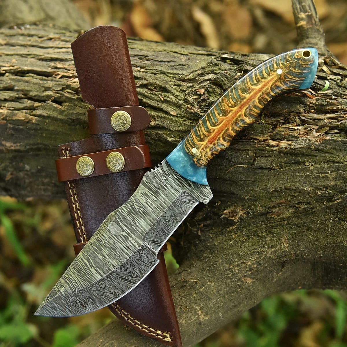 Hand-forged Damascus steel tracker knife with blue pinecone resin handle resting across a brown leather sheath on a mossy log