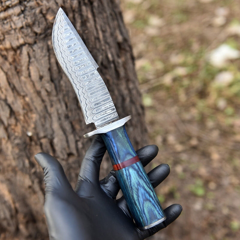 Hand-forged ladder pattern Damascus steel Bowie knife with a blue dyed wood handle and red spacer held in hand outdoors against a tree trunk.