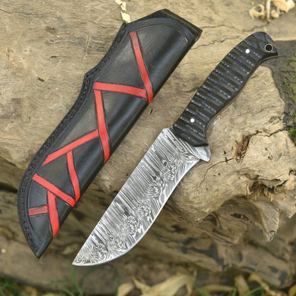 Hand-forged Damascus steel fixed blade knife with textured black G10 handle lying next to a black leather sheath with red geometric accents on wood log