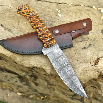 Hand-forged Damascus steel hunting knife with textured orange handle resting on a wooden log beside a basketweave brown leather sheath