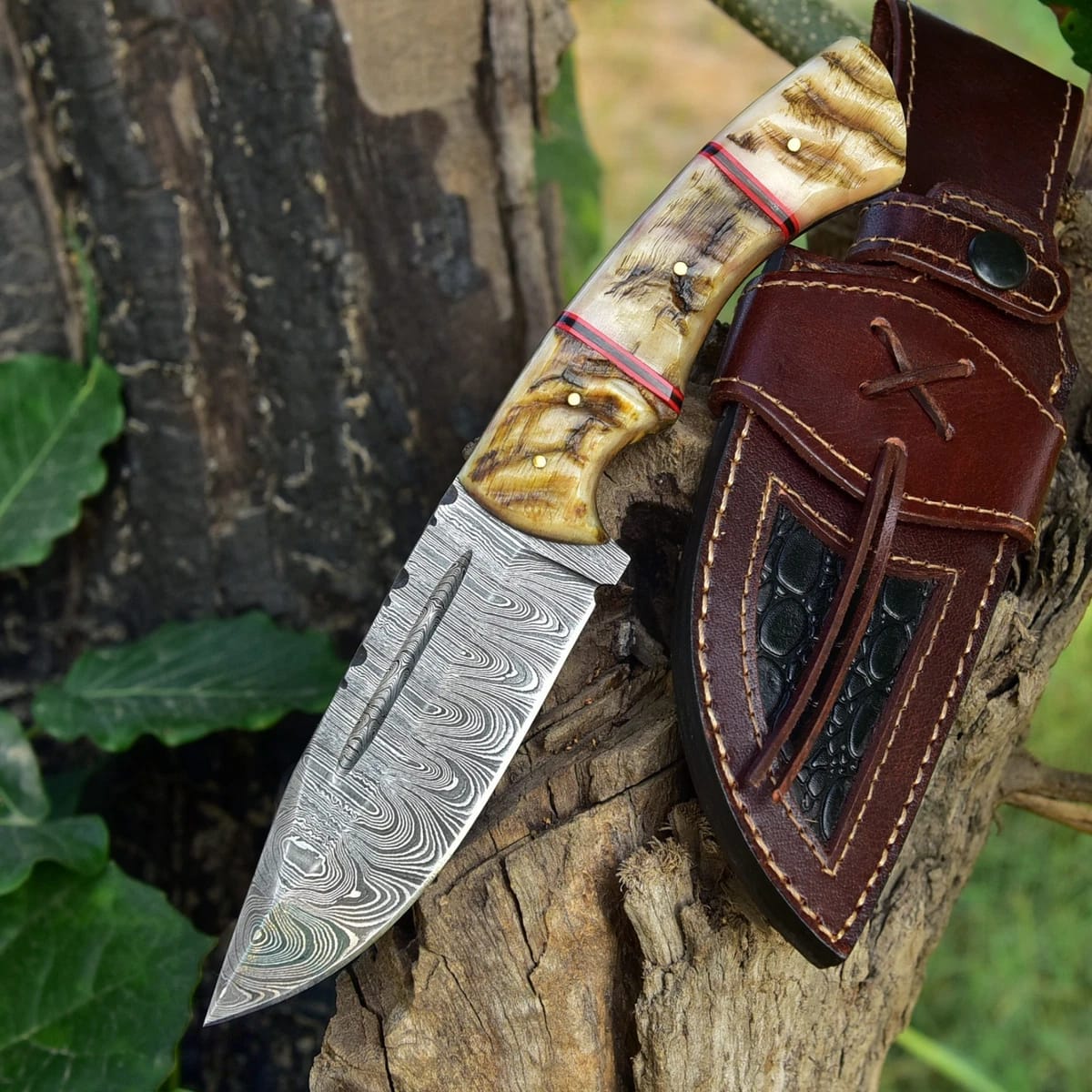 Hand-forged Damascus steel hunting knife with carved ram horn handle resting on a tree stump next to a brown leather sheath with dark inlay