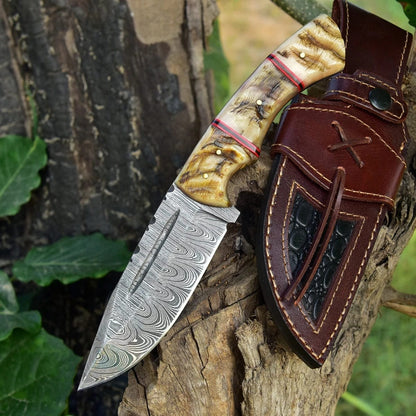 Hand-forged Damascus steel hunting knife with carved ram horn handle resting on a tree stump next to a brown leather sheath with dark inlay