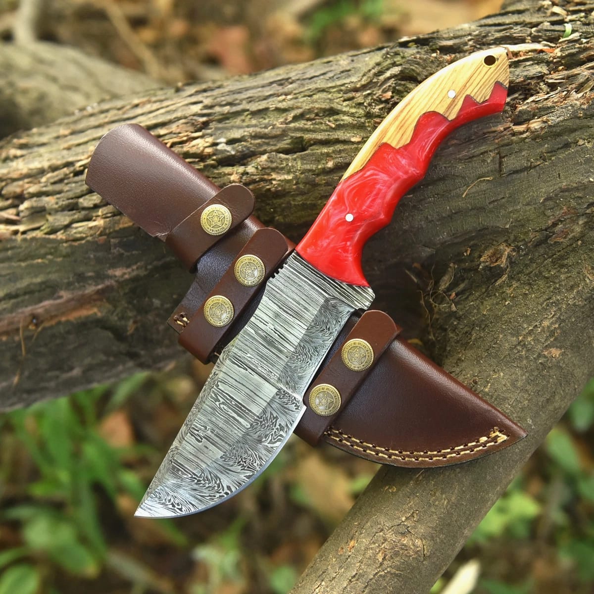 Hand-forged Damascus steel tracker knife with red resin and wood handle resting on a tree log next to a brown leather sheath with star snaps