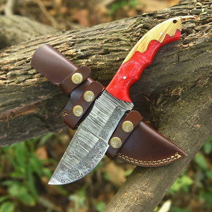 Hand-forged Damascus steel tracker knife with red resin and wood handle resting on a tree log next to a brown leather sheath with star snaps