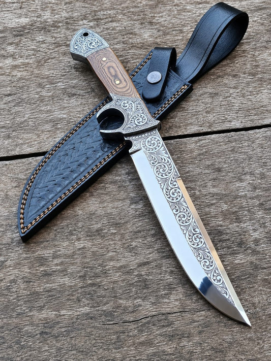 Hand-forged fixed blade Bowie knife with detailed scrollwork engraving and natural wood handle resting on weathered timber beside a black leather sheath.